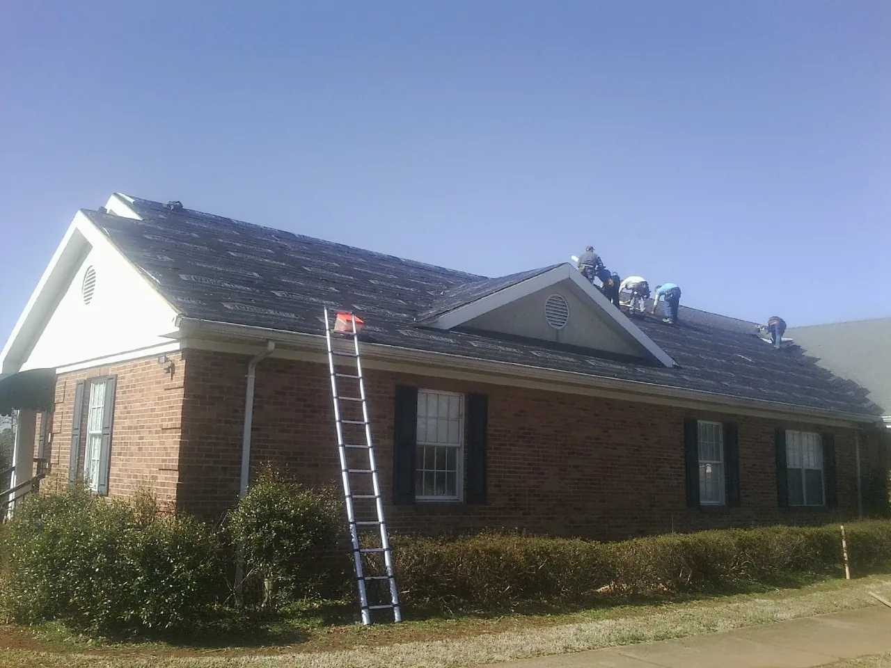 Crew tearing shingles off a brick ranch roof with a ladder set — Emergency Roof Repair work by our crew in Losantville
