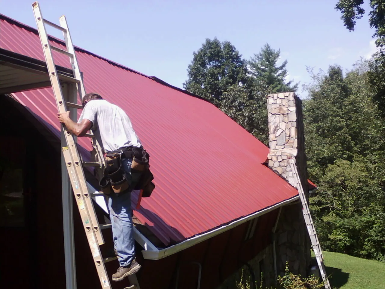 Roofer on a ladder beside a red metal roof and stone chimney — Soffit & Fascia Repair by Roofing Brothers in Losantville